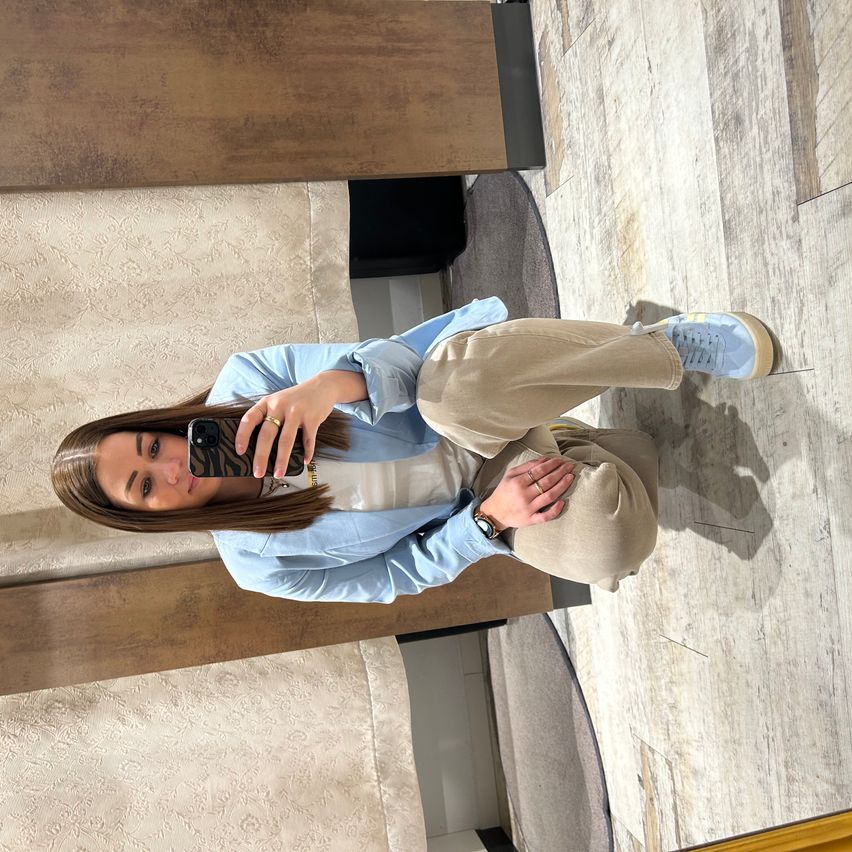 A woman is taking a selfie in a room with wooden floors and furniture. She wears a blue jacket, beige pants, and sneakers.