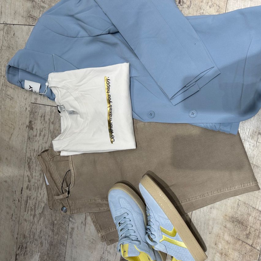 A blue blazer, a white t-shirt, beige trousers, and a pair of light blue sneakers with yellow accents are laid out on a wooden floor.