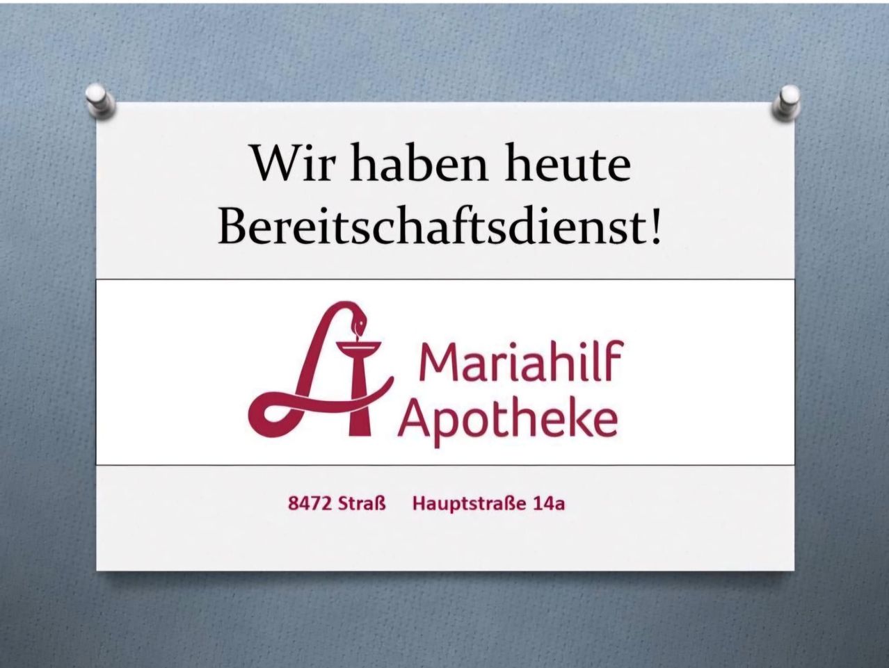 Ein weißes Schild mit blauem Hintergrund. Es steht 'Wir haben heute Bereitschaftsdienst!' und hat ein Logo mit einer roten Schlange und 'Mariahilf Apotheke'. Die Adresse lautet '8472 StraB HauptstraBe 14a'.