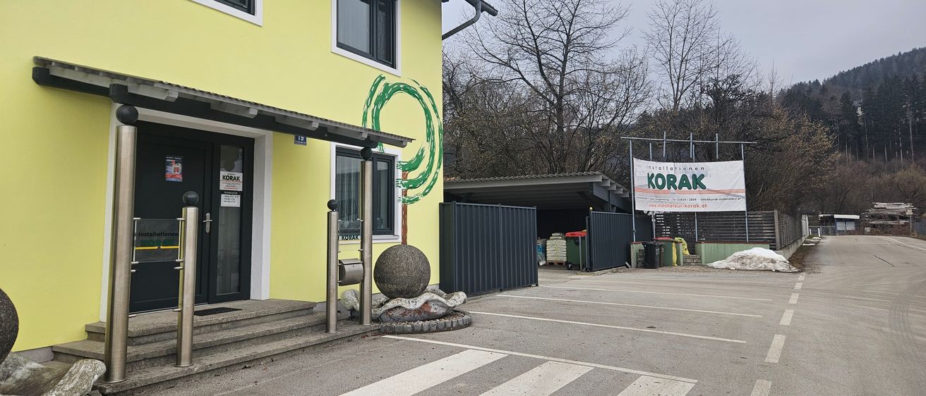 Der Eingang eines gelben Gebäudes mit einem grünen Spiraldesign an der Wand. Eine Baumskulptur und ein Schild mit der Aufschrift KORAK stehen vor dem Gebäude. Dahinter befinden sich mehrere Bäume und ein Banner mit dem Wort KORAK.