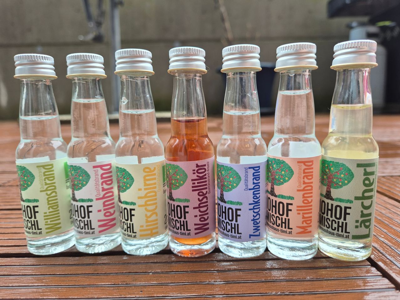 Acht kleine Flaschen mit Flüssigkeit, beschriftet mit Baumbildern und Text, sind auf einem Holztisch angeordnet. Die Etiketten enthalten die Worte 'HOF MUSCHL' und 'Weinbrand'.