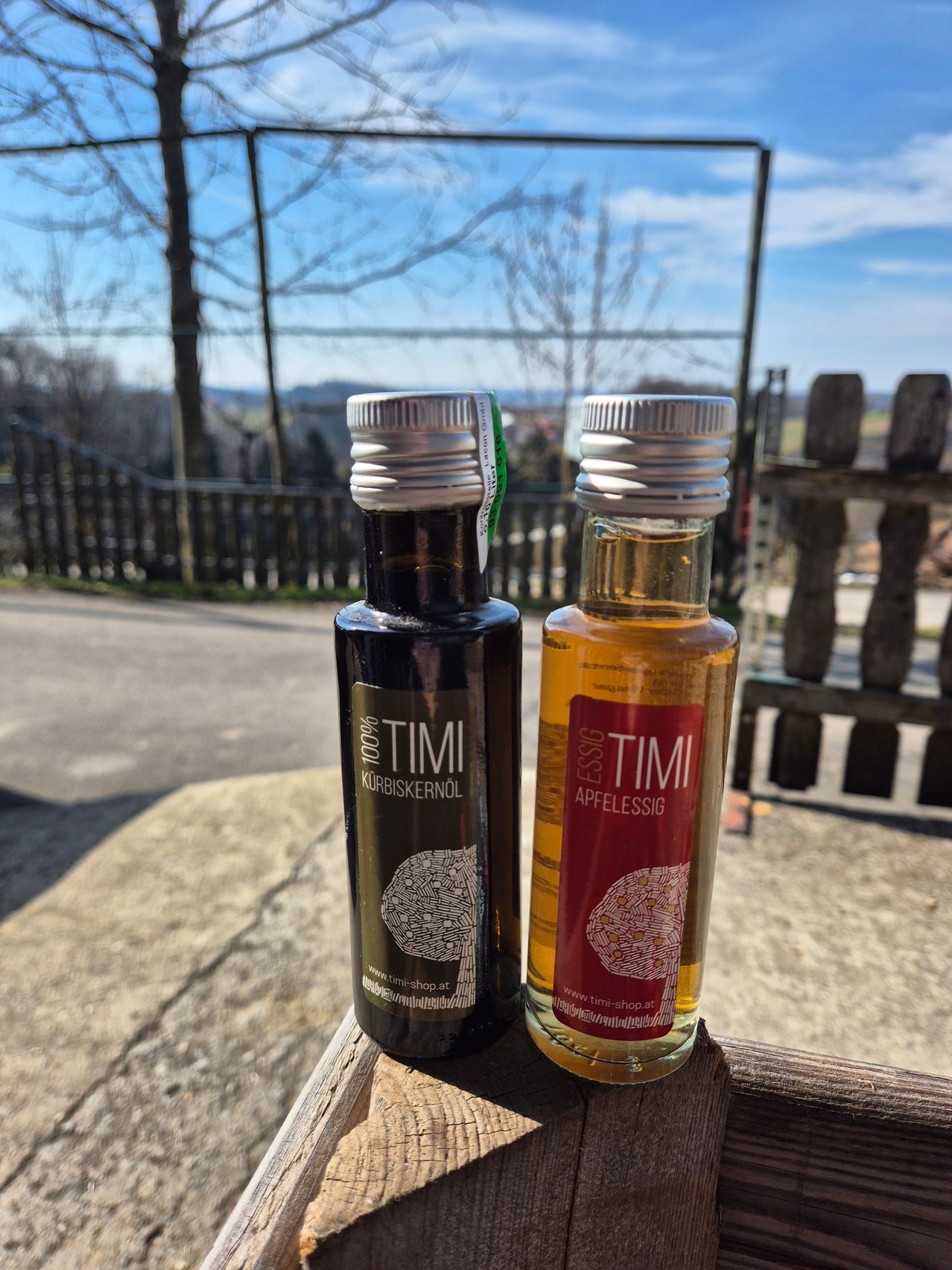 Two bottles of Timi, one labeled 'Kurbiskernol' and the other 'Apfelessig', are placed on a wooden surface outdoors near a wooden fence.