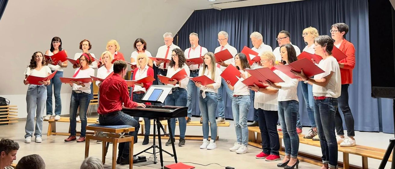 Ein Chor singt auf der Bühne mit einem Pianisten, der rote Bücher in der Hand hält. Das Publikum sitzt vor ihnen.