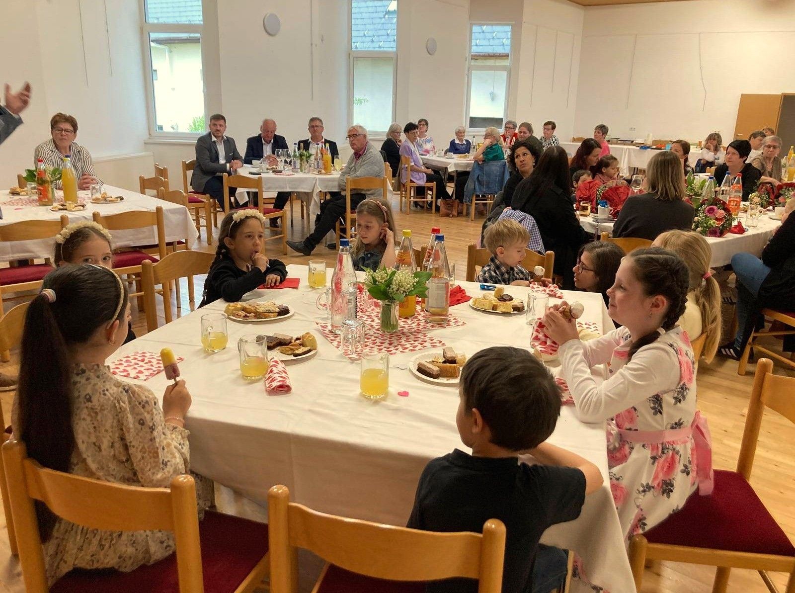 Eine Gruppe von Menschen, darunter Kinder, sitzt um einen Tisch herum, isst und trinkt. Der Tisch ist mit einer weißen Tischdecke bedeckt. Es gibt Teller mit Essen, Gläser mit Saft und eine Vase mit Blumen. Einige Menschen stehen und sitzen im Hintergrund auf Stühlen.