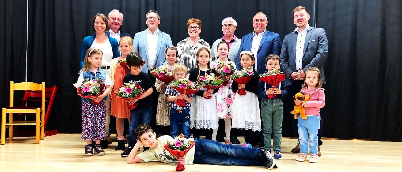 Eine Gruppe von Erwachsenen und Kindern auf der Bühne, einige halten Blumen und lächeln für ein Foto mit schwarzem Hintergrund und Holzboden.