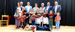 Eine Gruppe von Erwachsenen und Kindern auf der Bühne, einige halten Blumen und lächeln für ein Foto mit schwarzem Hintergrund und Holzboden.