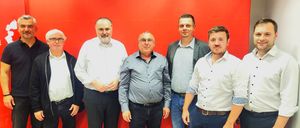 Vier Männer stehen lächelnd vor einer roten Wand. Der Mann auf der linken Seite hat ein weißes Hemd, schwarze Hose und einen Bart. Der zweite trägt ein graues Hemd und eine Brille. Der dritte trägt einen schwarzen Blazer über einem karierten Hemd. Der vierte trägt ein weißes Hemd und blaue Jeans.