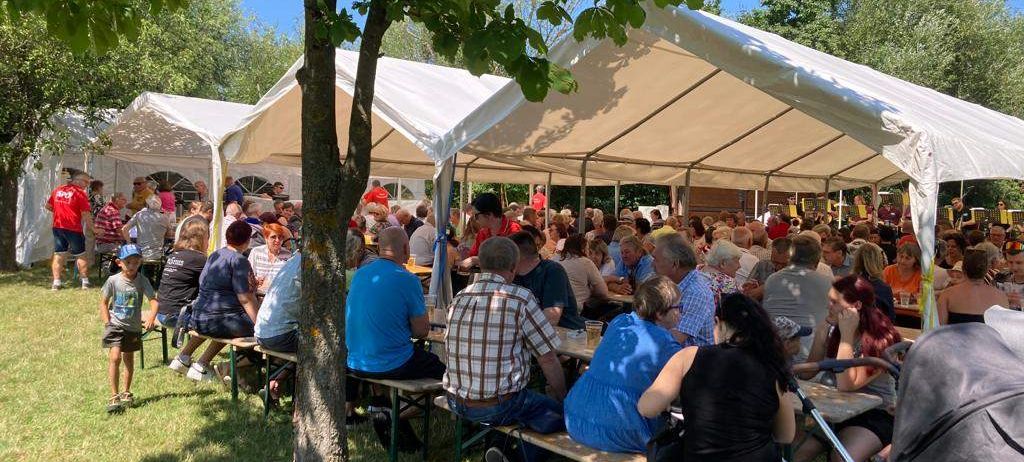 Eine Menschenmenge sitzt unter großen weißen Zelten an Tischen an einem sonnigen Tag und genießt Essen und Getränke. Einige tragen Hüte und Sonnenbrillen, und in der Nähe steht ein Kinderwagen.