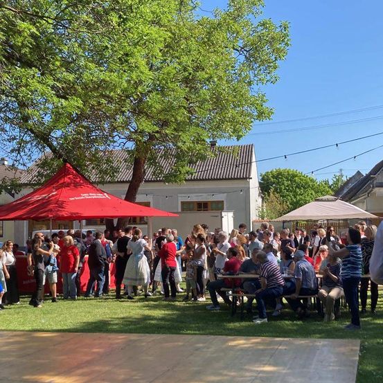 Eine Menschenmenge versammelt sich unter einem roten Zelt in einem Park. Einige sitzen an Tischen, während andere tanzen. Es gibt auch Regenschirme und ein Haus im Hintergrund.