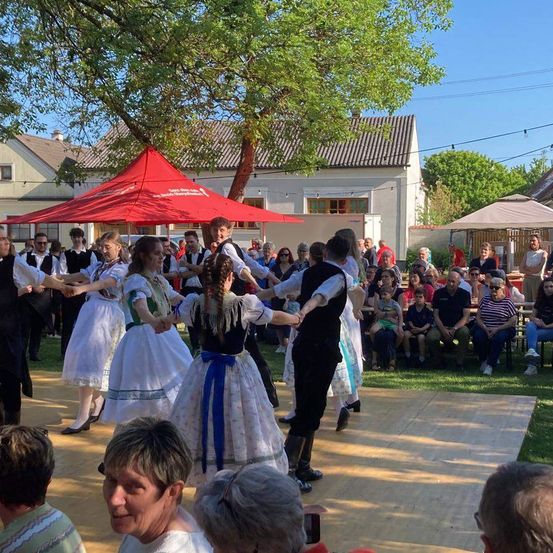 Eine Gruppe von Menschen tanzt im Freien. Sie tragen traditionelle Kleidung. Einige stehen im Kreis, andere sitzen auf dem Gras und schauen zu. Im Hintergrund befinden sich Häuser und Bäume.