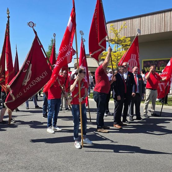 Eine Gruppe von Menschen in roten T-Shirts hält rote Fahnen und steht in einer Reihe auf einer Straße. Einige tragen Brillen und Uhren. Ein Mann vorne hält eine Fahne. Im Hintergrund befindet sich ein Gebäude mit einem gelben Banner.