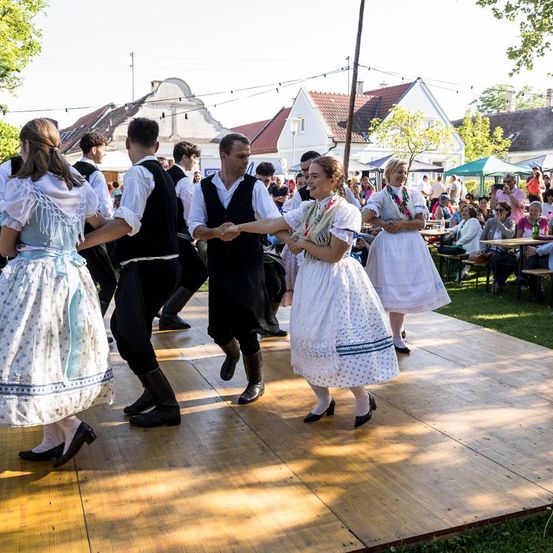 Eine Gruppe von Menschen in traditioneller Kleidung tanzt auf einer Holzbühne im Freien. Im Hintergrund befinden sich Häuser und Bäume.