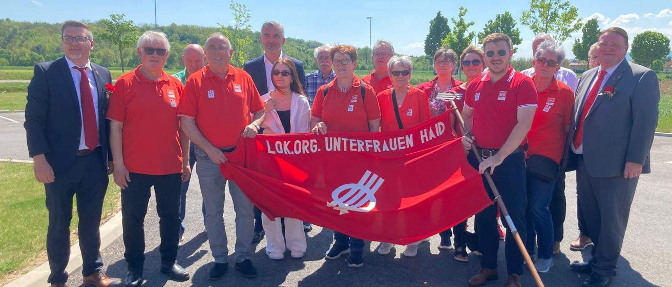 Eine Gruppe von Menschen in roten Hemden hält eine rote Fahne mit weißem Text. Sie stehen in einem sonnigen Außenbereich.