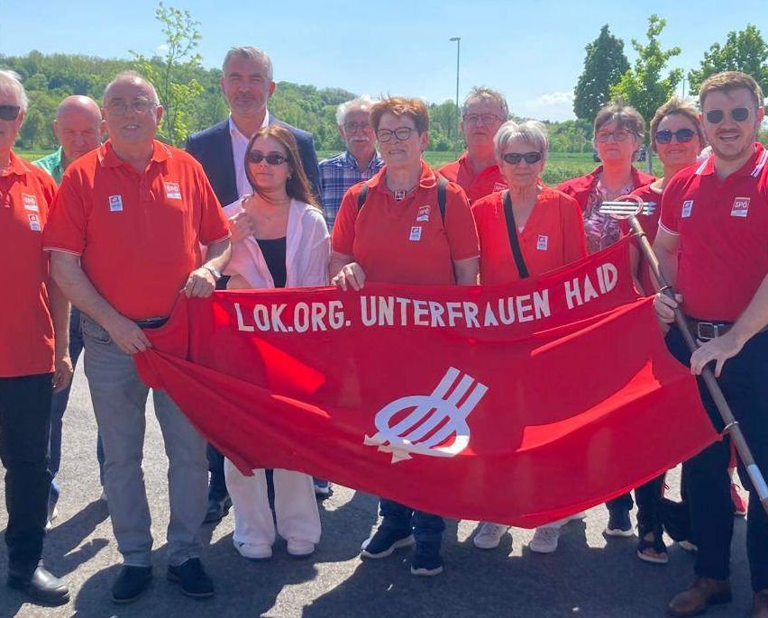 Eine Gruppe von Menschen in roten Hemden hält eine rote Fahne mit weißem Text. Sie stehen in einem sonnigen Außenbereich.