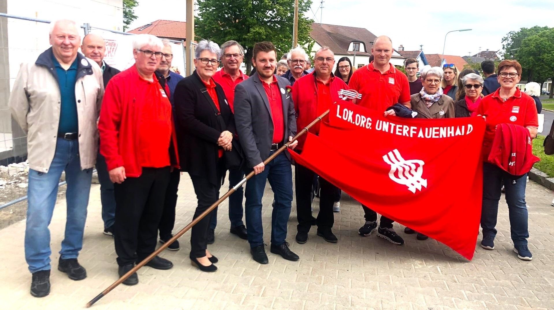 Eine Gruppe von Menschen in roten Hemden und Anzügen, die zusammenstehen. Ein Mann hält eine rote Fahne mit weißem Text. Sie stehen auf einem gepflasterten Weg.