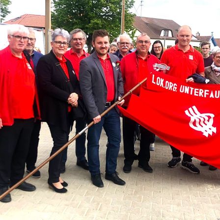 Eine Gruppe von Menschen in roten Shirts, die ein rotes Banner mit dem Text 'LOK.ORG UNTERFAUR' halten, steht vor einem Haus.