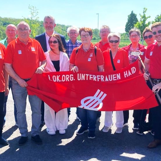 Eine Gruppe von Menschen, sowohl Männer als auch Frauen, in roten Shirts halten ein rotes Banner mit dem Text LOK.ORG. UNTERFRAUEN HAID. Sie stehen in einer Reihe.