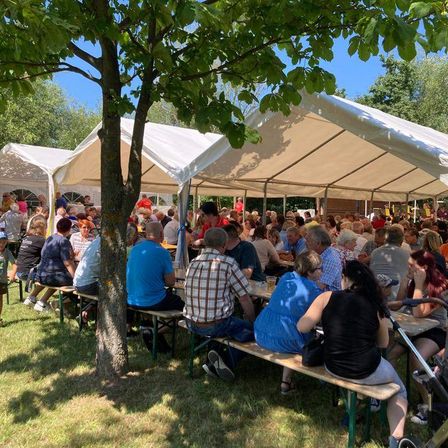 Eine große Gruppe von Menschen versammelt sich unter weißen Zelten in einem Außenbereich. Einige sitzen auf Bänken, während andere stehen. Es gibt Getränke auf den Tischen, und die Umgebung scheint eine Gemeinschaftsveranstaltung zu sein.