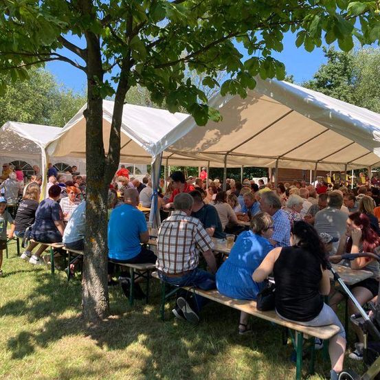Eine große Gruppe von Menschen versammelt sich unter weißen Zelten in einem Außenbereich. Einige sitzen auf Bänken, während andere stehen. Es gibt Getränke auf den Tischen, und die Umgebung scheint eine Gemeinschaftsveranstaltung zu sein.