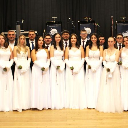 Ein Chor in weißen Kleidern posiert für ein Foto auf einer Bühne mit Musikinstrumenten und Bannern im Hintergrund.