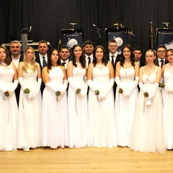 Ein Chor in weißen Kleidern posiert für ein Foto auf einer Bühne mit Musikinstrumenten und Bannern im Hintergrund.