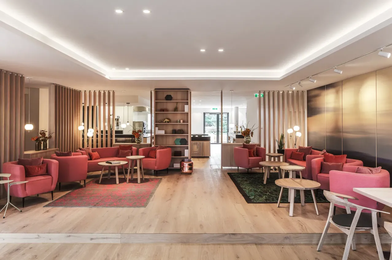 Ein Interieur mit Holzböden, einer hohen Decke und mehreren roten Stühlen um Tische mit roten Teppichen und Pflanzen. Ein großer Regalboden hält verschiedene Gegenstände und es gibt eine große Wand mit einem Bild.