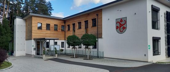 Die Vorderseite eines modernen Gebäudes mit Holzpaneelen. Zwei Bäume stehen davor und ein Logo an der Wand. Es gibt Glasfenster und ein Metallzaun an der Seite.