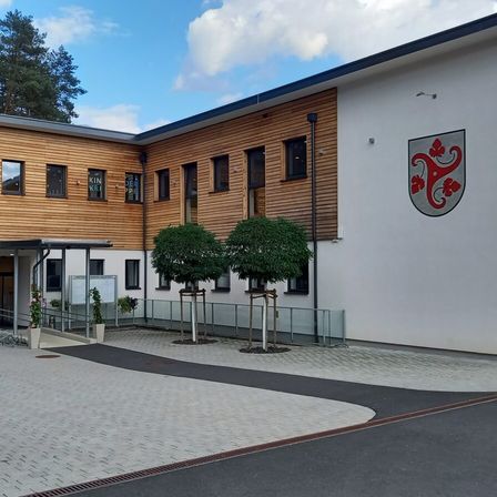 Das Äußere eines modernen Gebäudes mit Holzverkleidung, das ein großes Emblem an der Wand und mehrere Fenster aufweist. Zwei kleine Bäume stehen davor, mit Pflanzen und einem Weg, der zum Eingang führt.