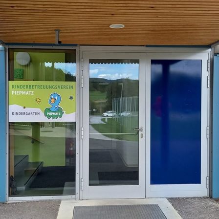 Der Eingang eines Kindergartens mit einer blauen Tür und einem Schild, das 'Kinderbetreuungsverein Piepmatz' steht. Drinnen ist eine grüne Wand mit einem Vogellogo.