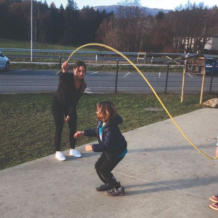 Zwei Mädchen spielen mit einem gelben Seil in einem Park, mit einem Zaun und einer Rasenfläche im Hintergrund.