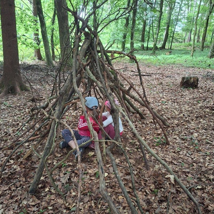 Zwei Kinder sitzen in einer improvisierten Hütte aus Ästen und Zweigen im Wald.