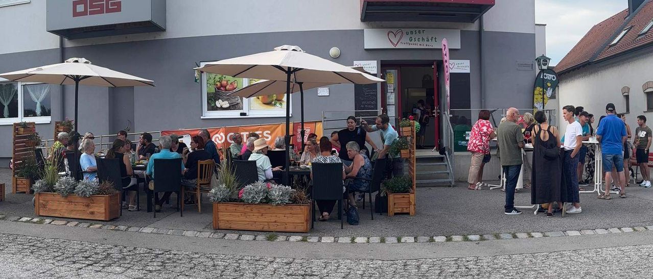 Mehrere Personen sitzen unter einem Regenschirm an Tischen vor einem Café. Der Eingang ist offen und eine Treppe führt hinauf. Bepflanzte Töpfe befinden sich auf beiden Seiten.