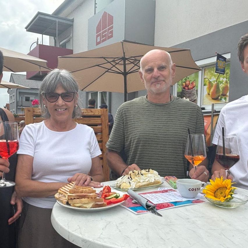 Drei Personen sitzen an einem Tisch im Freien und genießen Essen und Getränke. Eine Frau in Sonnenbrille und weißem T-Shirt hält ein Weinglas. Ein Mann in einem grünen T-Shirt hält eine Tasse. Ein anderer Mann in einem weißen T-Shirt lächelt.