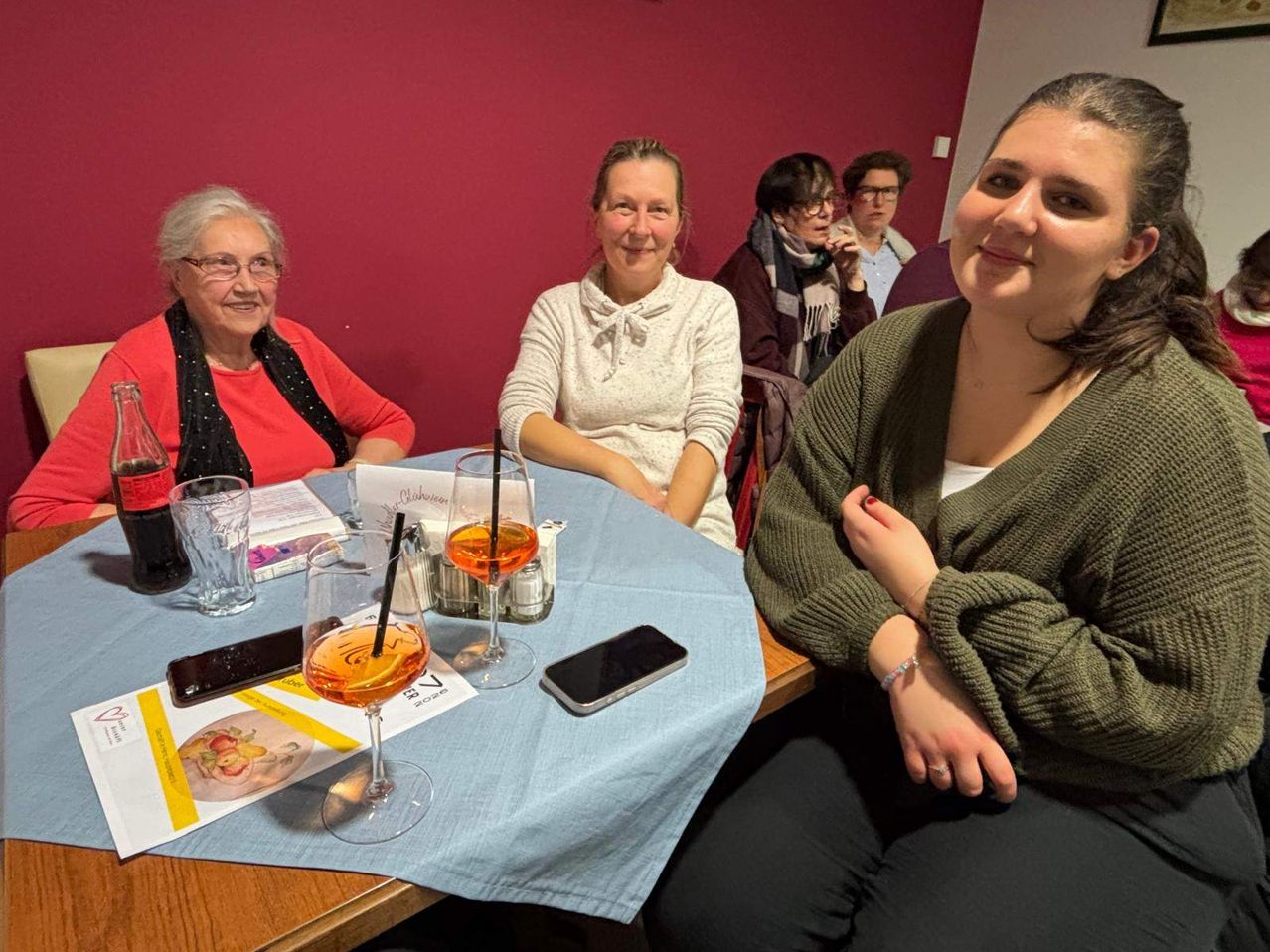 Eine Gruppe von Frauen sitzt um einen Tisch herum, lächelt und schaut in die Kamera. Eine Frau hat ihre Hände auf der Brust. Auf dem Tisch stehen Weingläser, ein Buch und Telefone.