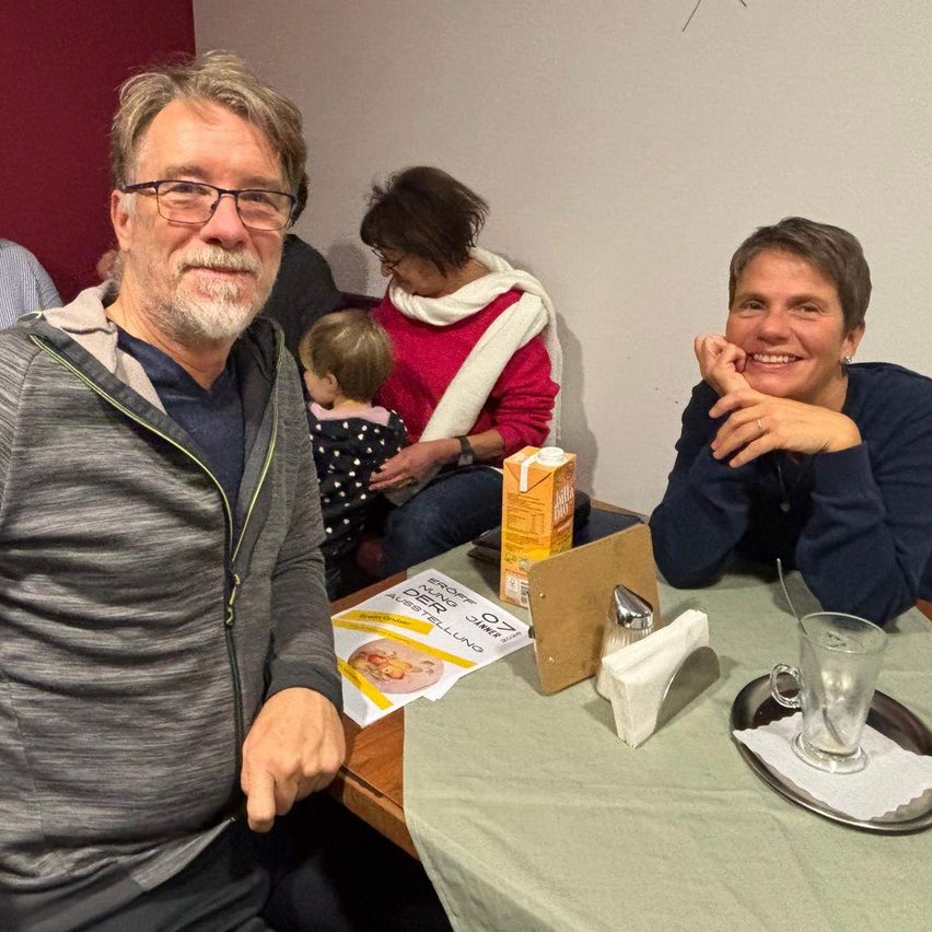 Drei Erwachsene und ein Kind sitzen lächelnd an einem Tisch. Eine Speisekarte und ein Glas Saft stehen auf dem Tisch. Hinter ihnen ist eine Wand zu sehen.