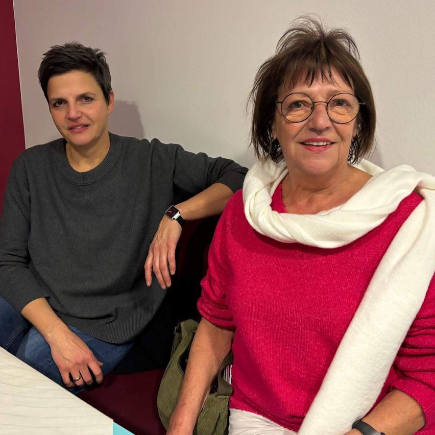 Zwei Frauen sitzen auf einer Couch, eine trägt ein graues Sweatshirt, die andere ein rotes Shirt mit einem weißen Schal. Sie lächeln beide, eine mit Brille, und sie sind in einem Raum mit einer weißen Wand.