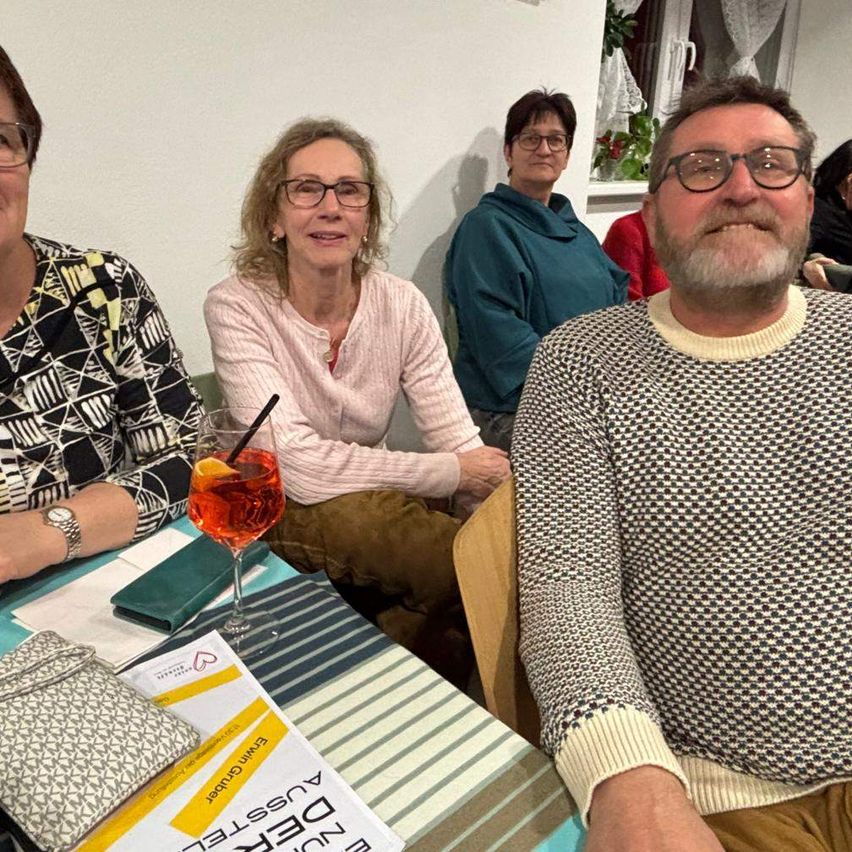 Eine Gruppe von Menschen sitzt um einen Tisch mit einem Glas Wein und lächelt für ein Foto. Die Frau in Pink trägt eine Brille und der Mann in Grau hat einen Bart.