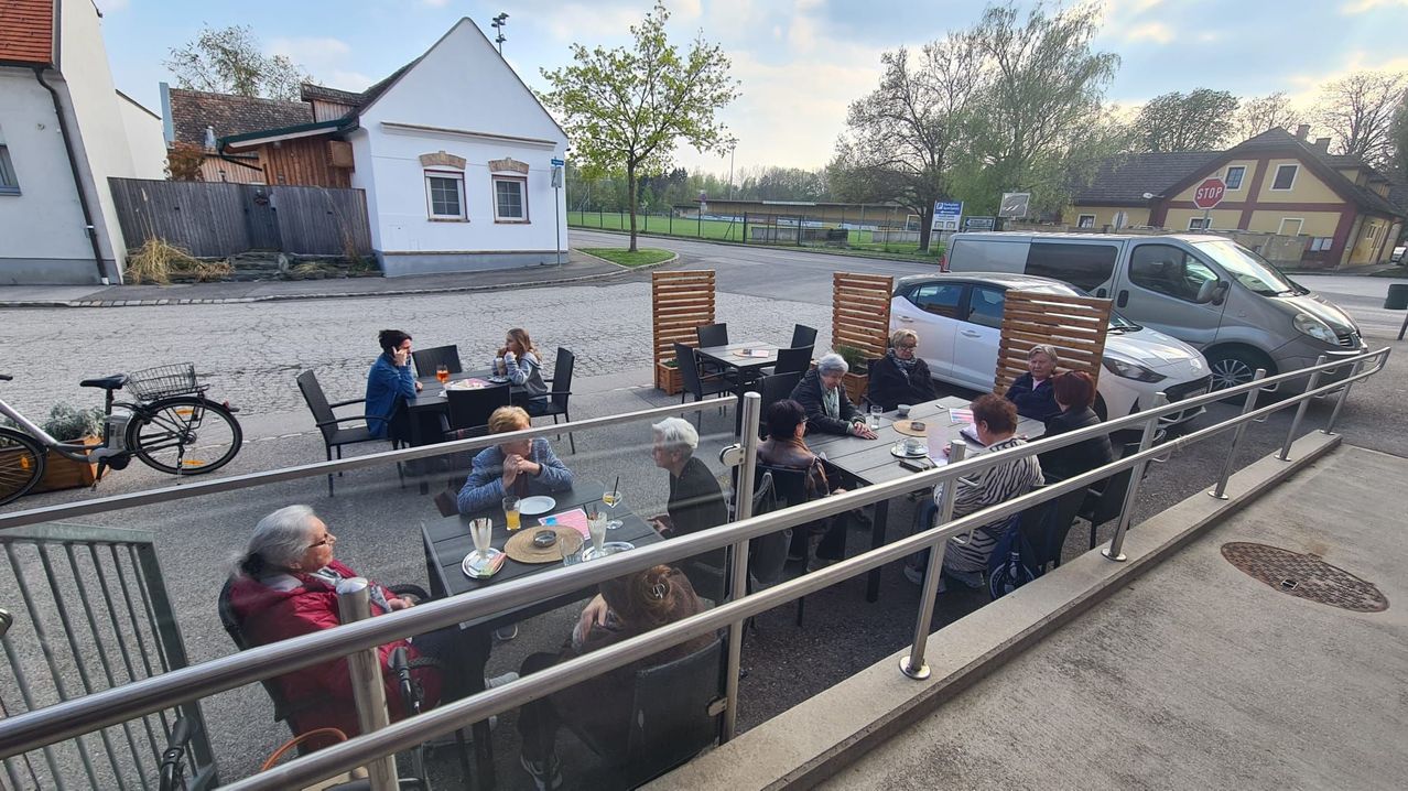 A group of elderly people is sitting around tables in an outdoor area with a glass barrier. There are chairs, a table, and a glass cup with straw on top. Behind them is a white van, and in the distance is a house with a tree in front.