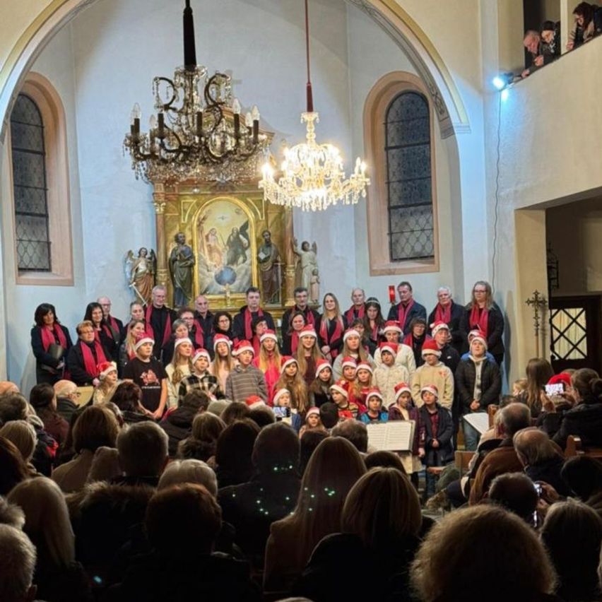 Ein Chor in Weihnachtsmützen tritt in einer Kirche auf. Das Publikum sitzt vor ihnen. Über dem Chor hängen zwei Kronleuchter.