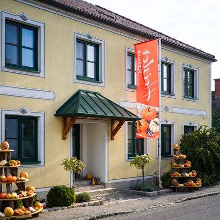 Ein Gebäude mit einem grünen Vordach und einer roten Fahne mit Kürbissen davor. Auch Kürbisse sind auf einem Ständer links neben dem Gebäude angeordnet.