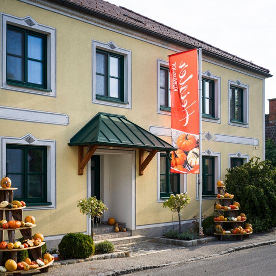 Ein Gebäude mit einem grünen Vordach und einer roten Fahne mit Kürbissen davor. Auch Kürbisse sind auf einem Ständer links neben dem Gebäude angeordnet.