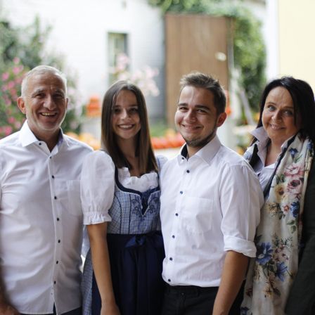 Vier lächelnde Erwachsene und eine junge Frau stehen dicht beieinander draußen, möglicherweise für ein Foto. Sie sind in weißen Hemden und Freizeitkleidung gekleidet. Im Hintergrund befinden sich ein Gebäude und Pflanzen.