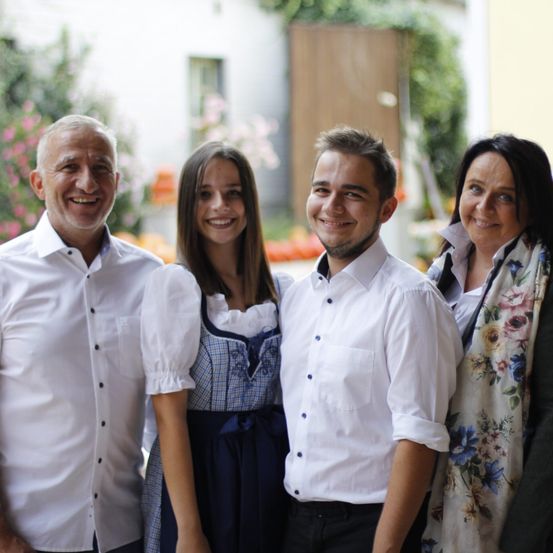 Vier lächelnde Erwachsene und eine junge Frau stehen dicht beieinander draußen, möglicherweise für ein Foto. Sie sind in weißen Hemden und Freizeitkleidung gekleidet. Im Hintergrund befinden sich ein Gebäude und Pflanzen.