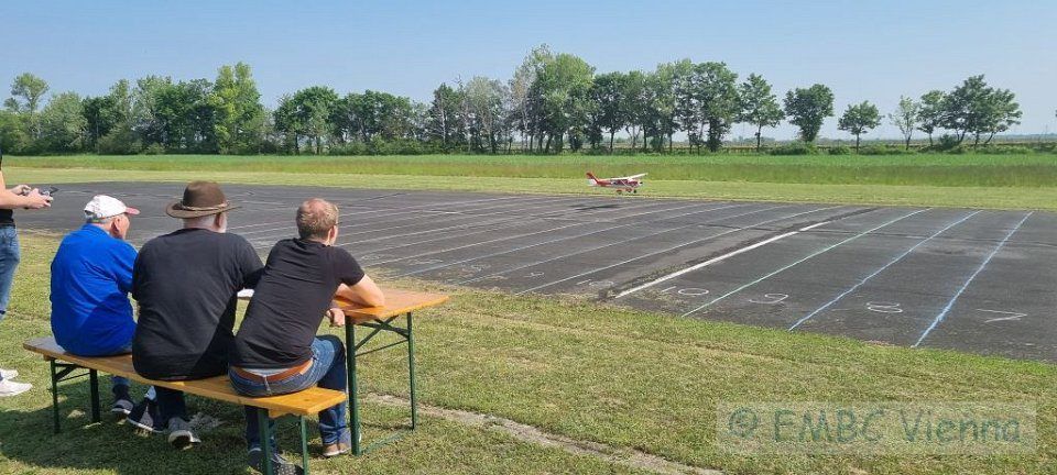 Drei Männer sitzen auf einer Bank an einem Flughafen und beobachten, wie ein kleines rotes Flugzeug von der Startbahn abhebt.