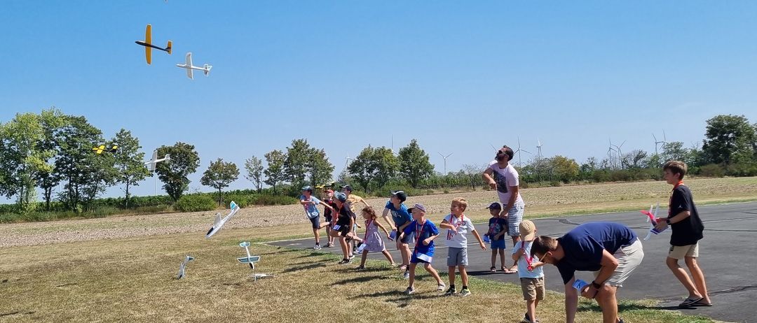 Mehrere Kinder und Erwachsene sind im Freien und fliegen oder beobachten ferngesteuerte Flugzeuge. Einige sind auf dem Gras, andere auf dem Asphalt. Bäume und Windturbinen sind im Hintergrund.