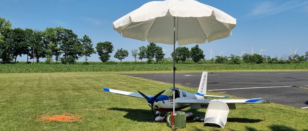 Ein kleines Flugzeug ist auf einem Grasfeld geparkt, mit einem großen weißen Regenschirm, der Schatten spendet. In der Nähe steht ein grüner Container auf dem Boden. Im Hintergrund sind Bäume und Windräder zu sehen.