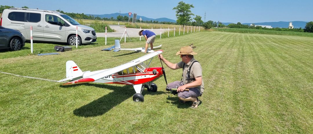 Zwei Männer bereiten ein rotes und weißes Modellflugzeug auf einem Grasfeld vor. Einer kniet und hält den Propeller, während der andere die Flügel justiert. Ein Auto und ein dreieckiges Schild sind im Hintergrund zu sehen.