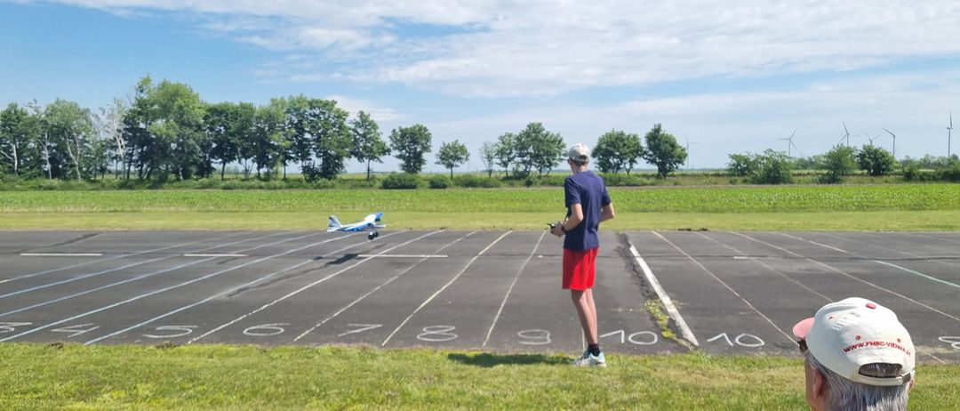 Eine Person mit Mütze und Turnschuhen steht auf einem Grasfeld und schaut auf ein ferngesteuertes Flugzeug, das auf einer markierten Startbahn fliegt.