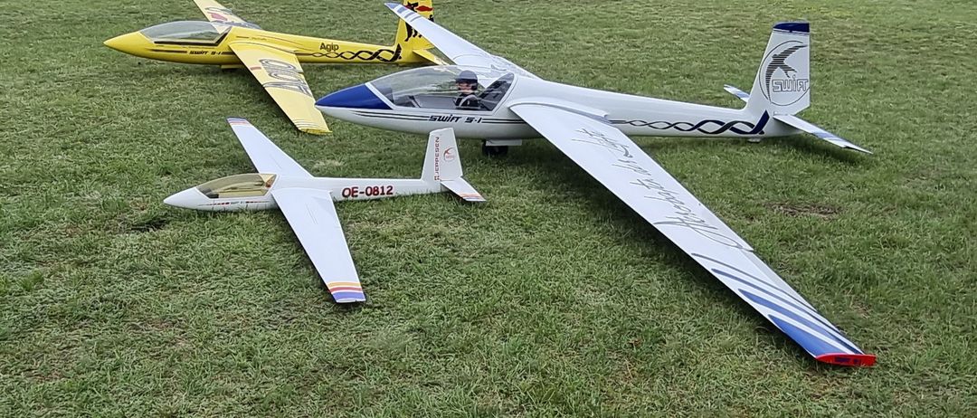 Drei Modellflugzeuge stehen auf einem Grasfeld. Das größte ist weiß mit einer blauen Kabine. Ein Pilot sitzt im Inneren. Die anderen beiden sind gelb und grau.