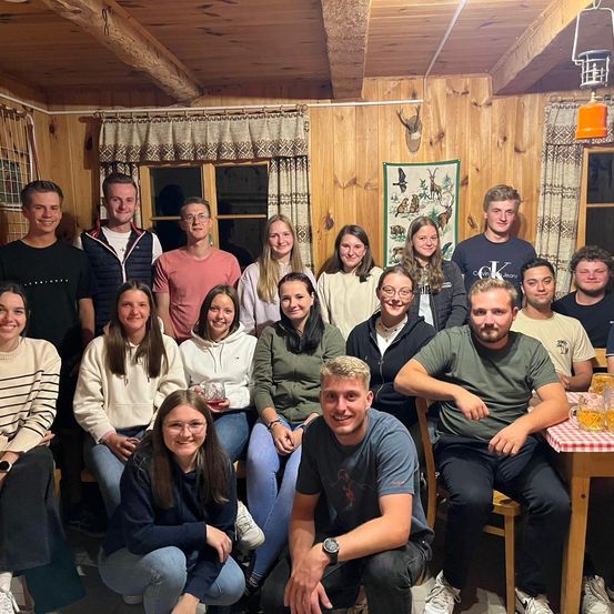 Eine Gruppe von Menschen posiert für ein Foto in einer Hütte mit Holzwänden und einem Fenster. Sie sitzen und stehen um einen Tisch mit Gläsern herum.